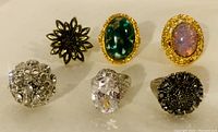 View of six ladies rings with stones and crystals arranged in two rows on white surface.