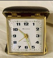 Front view of the vintage Bulova travel alarm clock in the open protective case, showing the clock face and hands, with the embossed brand name on the dial.