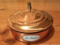 Top-down view of shiny copper lid with brass thumb lift knob, showing reflective surface and wooden table beneath.