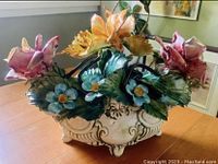 Front angle showing the full flower basket with colorful pink, yellow, and blue flowers and ornate bisque porcelain base on a table.