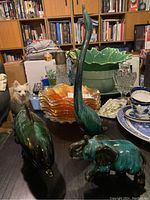 Photo showing all three ceramic figurines - green glossy heron, elephant, and dolphin on a table with other household items and blue dishware in background.