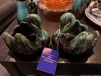 Pair of Blue Mountain Pottery swans shown side-by-side on a table with a tag attached to one swan labeled 'The Blue Mountain Collection'