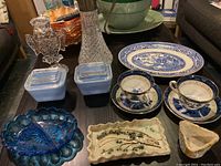 Display of all items on a table including Blue Willow platter, tea sets, Pyrex butter containers, crystal and silver decanter, blue pickle tray, ceramic butter dish, and crystal owl candle holder.