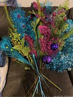 Assorted multicolored peacock themed Christmas picks in chartreuse, turquoise, and magenta with a turquoise tinsel wreath visible in the background.
