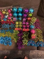 Photo shows multiple boxes of Christmas ball ornaments in blue, purple, green, and pink colors, along with loose star, snowflake, poinsettia, and peacock-shaped ornaments spread out on a sofa.