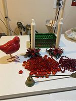 Lot of red Christmas holiday items displayed on a white table including a red bird figurine, two red candle holders with white taper candles, red glass beads, red garland beads, and a green trinket box.