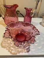 Large cranberry glass bowl with ruffled white rim and two cranberry glass pitchers behind it