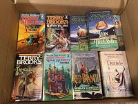 Top-down view of all 9 softcover novels arranged in a cardboard box showing cover artwork and titles.