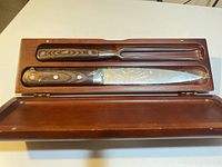 Meat carving knife and fork with rosewood handles inside a rosewood case, fully visible showing wood grain and metal blades/prongs.