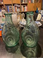 Two translucent green glass vases shaped like nautilus shells with spiral ridged detailing, sitting side by side on a wooden table