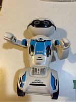 Full front view of a white robot toy with black and blue accents, arms raised outward, showing its head, eyes, and torso details including 'SBS' marking.