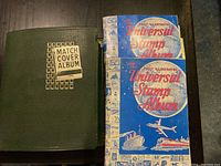 Two green and blue albums on a surface: green leatherette Match Cover Album with gold lettering and two blue Universal Stamp Albums with illustrated covers.