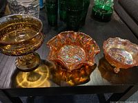 Three amber carnival glass bowls displayed on a dark table, showing iridescent shine and detailed designs.
