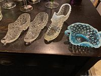 View of all four items: two clear pressed glass shoes, one white hobnail glass shoe, and one blue hobnail glass bowl on dark surface.
