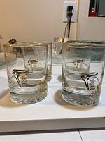 Set of four clear whiskey glasses with gold rim and Olympic torch design arranged on a white surface.