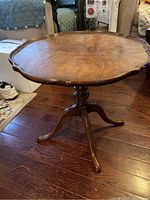 Full view of antique round wooden side table with pedestal base and scalloped edges standing on wood floor.