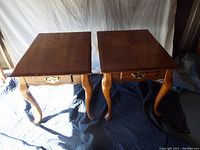 Pair of two wooden end tables on padded blanket with white backdrop, showing tops and front drawers with brass pull and curved legs.