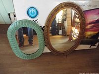 Large ornate oval mirror and smaller green wicker bordered oval mirror side by side on floor