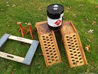 Pair of rusty yellow metal car ramps, two small red metal jack stands, blue cushioned dolly, and black metal drum with warning label on grass.
