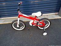 Side view of red and white child's bike with training wheels on pavement in front of blue rolling door.