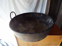Side view of large blackened metal bowl with two metal handles, showing patina and wear.