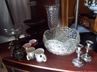 Various glassware items including tall crystal vase, punch bowl, large glasses, coffee cups, and metal candlesticks arranged on a wooden surface.