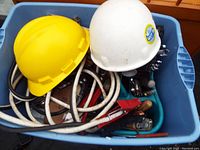 Blue plastic bin containing yellow and white hardhats, jumper cables, and various hand tools such as pliers, wrenches, and a hammer.