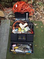 Photo showing black toolbox filled with assorted hand tools and hardware, alongside several coiled orange extension cords on outdoor ground.