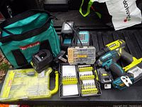 Makita cordless drill with battery, Ryobi battery charger, multiple drill bit sets in yellow and clear cases, and Makita soft bag on a truckbed tailgate.