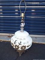 Full view of the lamp showing the white glass base with metal floral details and the metal base stand with lamp socket and harp.