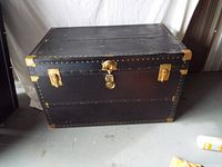 Front view of black rectangular storage chest with metal corner reinforcements, latch, lock and side clasps on concrete floor.