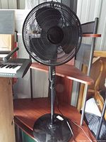 Black upright pedestal fan with round base, black mesh grille, and three blades shown on wooden table.