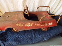 Side view of vintage red pedal car showing rust, worn decal, metal wheels, and steering wheel