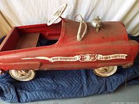 Side view of red metal pedal car showing fire department decal, white rusty wheels, metal steering wheel, and fire bell mounted on hood.