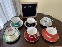 Full view of all six teacup and saucer pairs arranged around a sign showing lot number on a wood table.