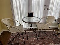 Full view of bistro table and two chairs positioned in front of window with sheer curtains. Showing front and side profiles of chairs and round glass tabletop.