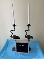 Pair of dark brown metal crane-shaped candlesticks holding taper candles, standing on a blue cloth with 'LOT 98' sign.