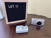 Canon PowerShot SX240 HS digital camera and battery charger on a wooden table seen front and with a black felt board reading LOT 11