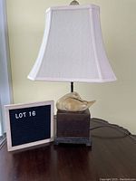 Table lamp with square base and ceramic duck on a wooden surface next to a sign showing Lot 16.