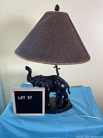 Front view of black elephant table lamp with faux wicker shade and power cord visible.