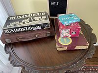 Games lot displayed on a wooden table including Rummikub, Rook, Scrabble, and Dominoes boxes showing wear and vintage condition.
