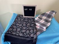 Both men's scarves displayed on blue surface with LOT 112 sign. One scarf is blue Paisley tube style, the other is brown plaid fringed.