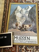 Full view of framed mountain scene poster with cows and text including 'Murren 1650 M.U.M. Bergbahn Funiculaire Suisse Berner-Oberland Switzerland' lying on patterned carpet.