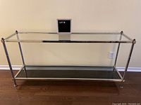 Front view of the sofa table showing clear glass top shelf, smoky glass bottom shelf, and shiny chrome frame on hardwood floor against beige wall