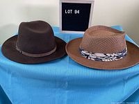 Two men's hats on blue cloth, one dark brown felt and one brown straw with floral band, shown side by side with 'LOT 94' sign.