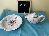 Photo shows white porcelain teapot and a decorative plate with floral and lattice design on a turquoise cloth.