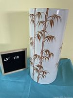 Front view of tall white ceramic umbrella stand featuring brown bamboo design, set on blue cloth with lot number board.