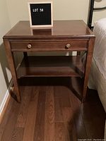 Front view of wooden night table with drawer and lower shelf on hardwood floor beside bed.