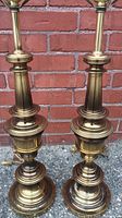 Pair of tall, classic turned brass lamp bases sitting against a brick wall, no shade attached, showing overall shape and brass finish.