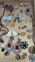 Top-down view of assorted costume jewelry on wooden tray, showing various earrings, rings, necklaces, and brooches with colorful stones and metallic finishes.
